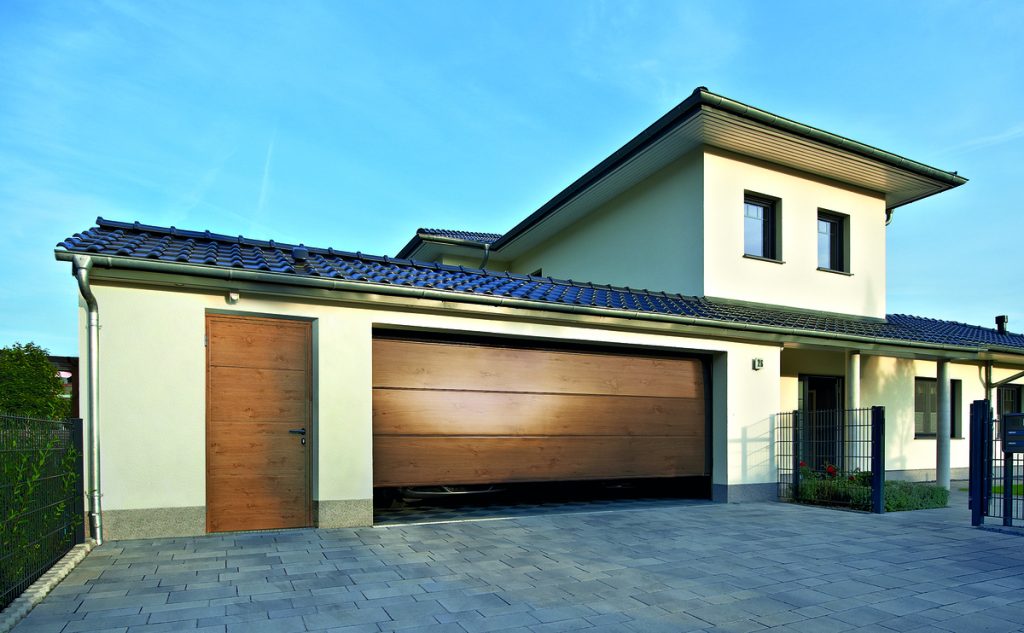 A contemporary cream-colored house with a tiled roof and attached garage. The garage has wooden horizontal-paneled doors that are partially open. There's a matching wooden side door next to the main garage. The driveway is made of large rectangular stone pavers, and the property is bordered by a metal fence. The architectural style blends traditional elements with modern design.