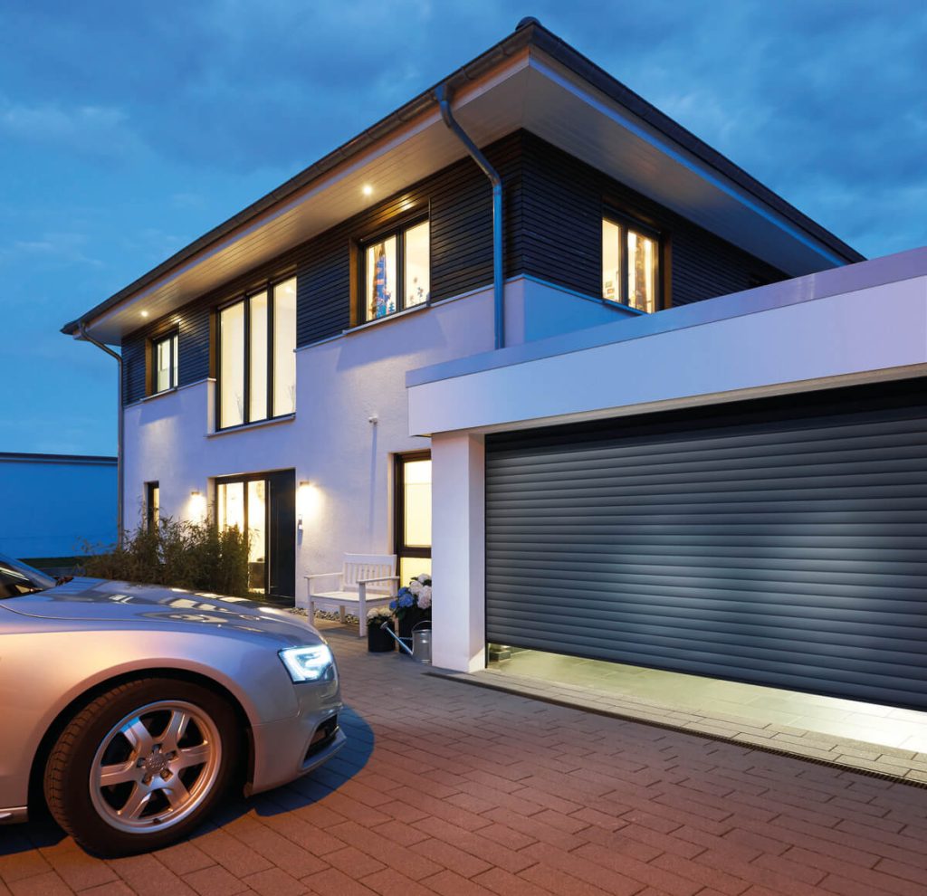 A modern two-story house photographed at dusk with lights on inside. The upper portion features dark horizontal siding and large windows, while the lower portion is white with a dark gray garage door. A car with headlights on is parked in the brick-paved driveway. The house has a flat roof design with overhanging eaves and exterior wall lighting creating a warm ambiance.