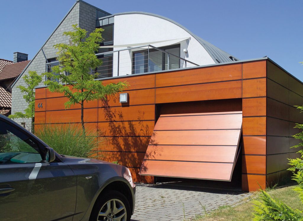 A unique modern house with a curved metal roof section and an attached wooden garage. The garage has a horizontal-paneled door that swings outward like a ramp. The garage exterior is clad in warm orange-toned wood panels arranged horizontally. The main house features gray stone or concrete blocks with glass balustrades on an upper terrace. A dark colored car is partially visible in the foreground, and young trees provide landscaping elements.
